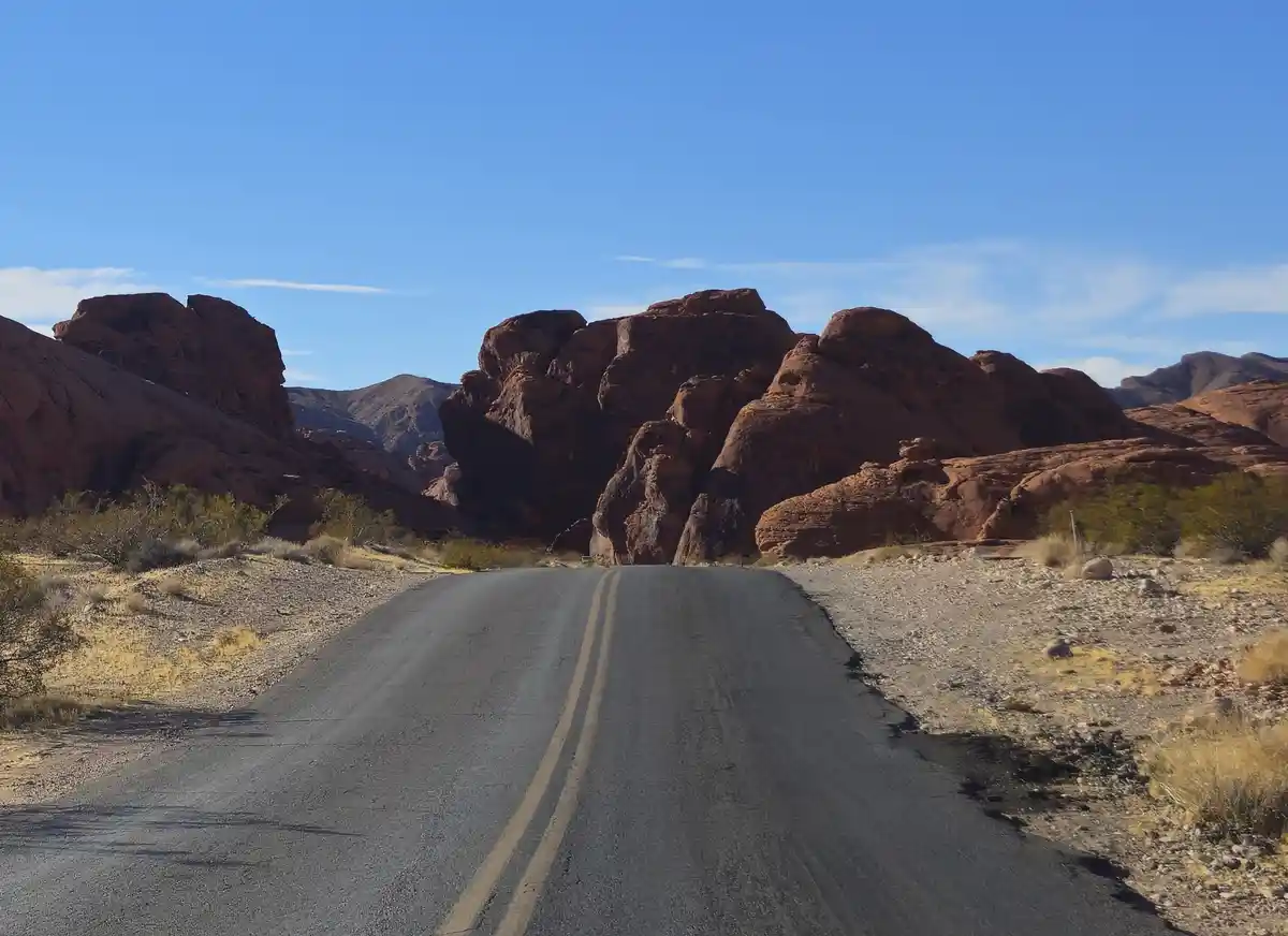 A straight asphalt road with double yellow lines stretches toward large red rock formations under a clear blue sky. Sparse…