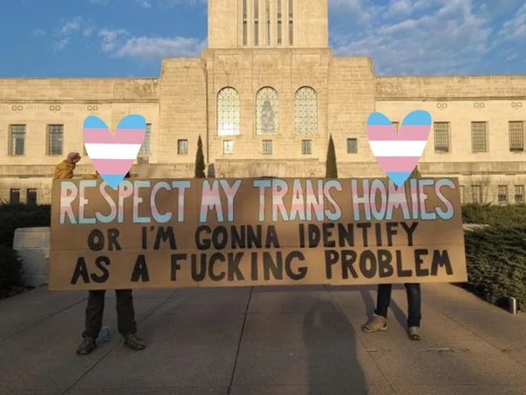 Two people hold a cardboard protest sign outdoors in front of a large stone building. The sign reads 'RESPECT MY TRANS…
