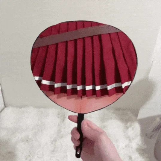 A hand holds a round fan with a red pleated skirt design on one side and an anime girl with red hair and an eyepatch on the…