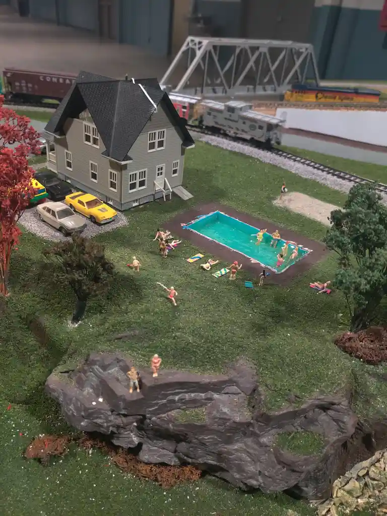 A detailed model train diorama features a miniature house, cars, and a swimming pool with tiny figurines enjoying a summer…