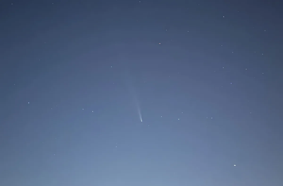 A night sky image shows a bright comet with a long, diffuse tail streaking diagonally across a dark blue sky dotted with…