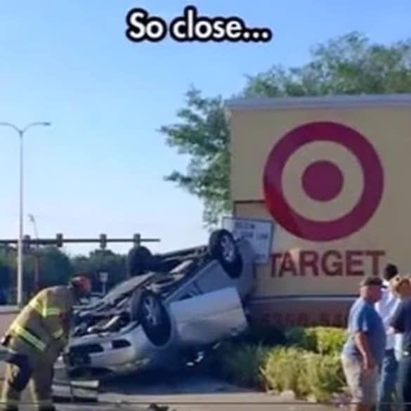 A silver car is flipped upside down and crashed into the side of a Target store sign. A firefighter in yellow gear is…