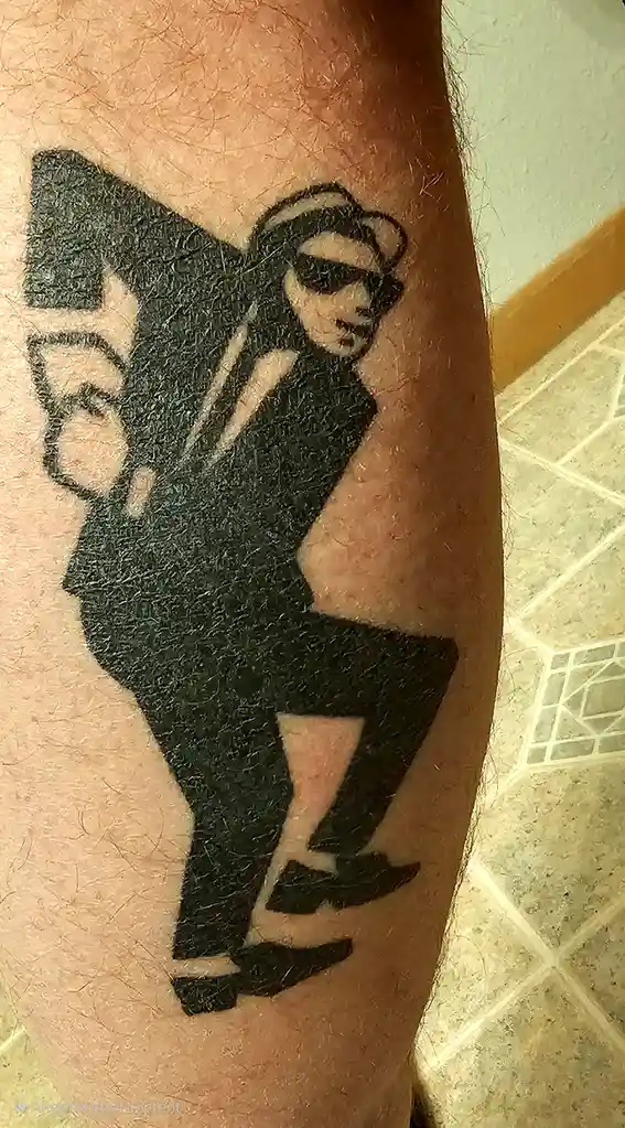 A close-up photo shows a black ink tattoo on a person's arm, depicting a stylized man in a suit, sunglasses, and a fedora…