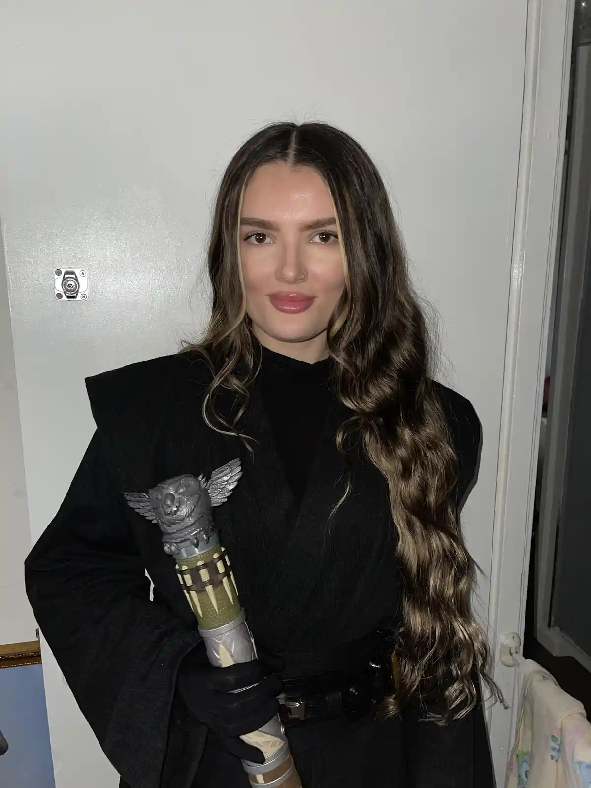 A woman with long brown hair and a nose ring wears a black robe and black gloves, holding a detailed lightsaber hilt with a…