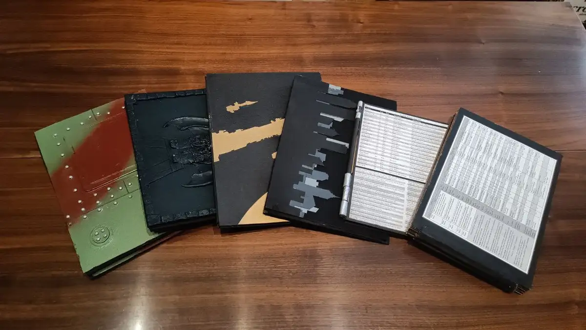 A row of five custom-made, heavily modified, and artistically painted tabletop wargaming codex binders lie open on a wooden…