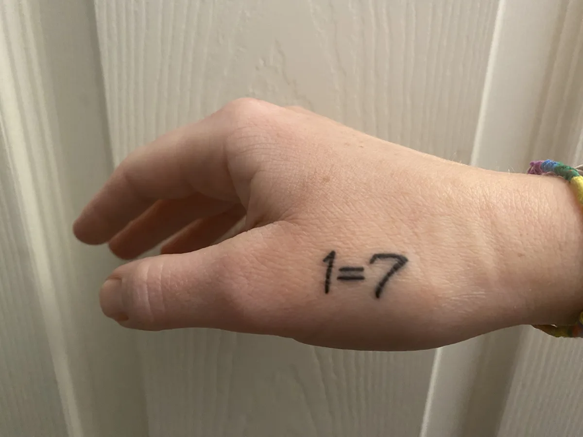 A close-up photo shows a person's left hand with the handwritten text '1=7' written in black marker on the palm. The hand is…