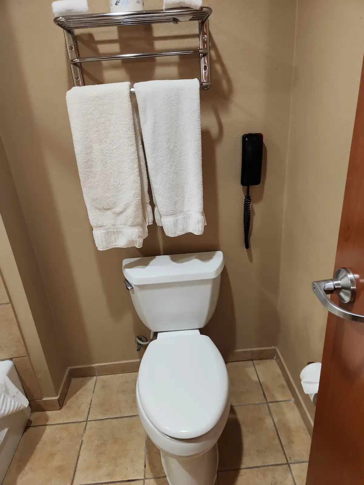 A bathroom scene featuring a white toilet with the lid down, a chrome towel rack above holding two white towels, and a…