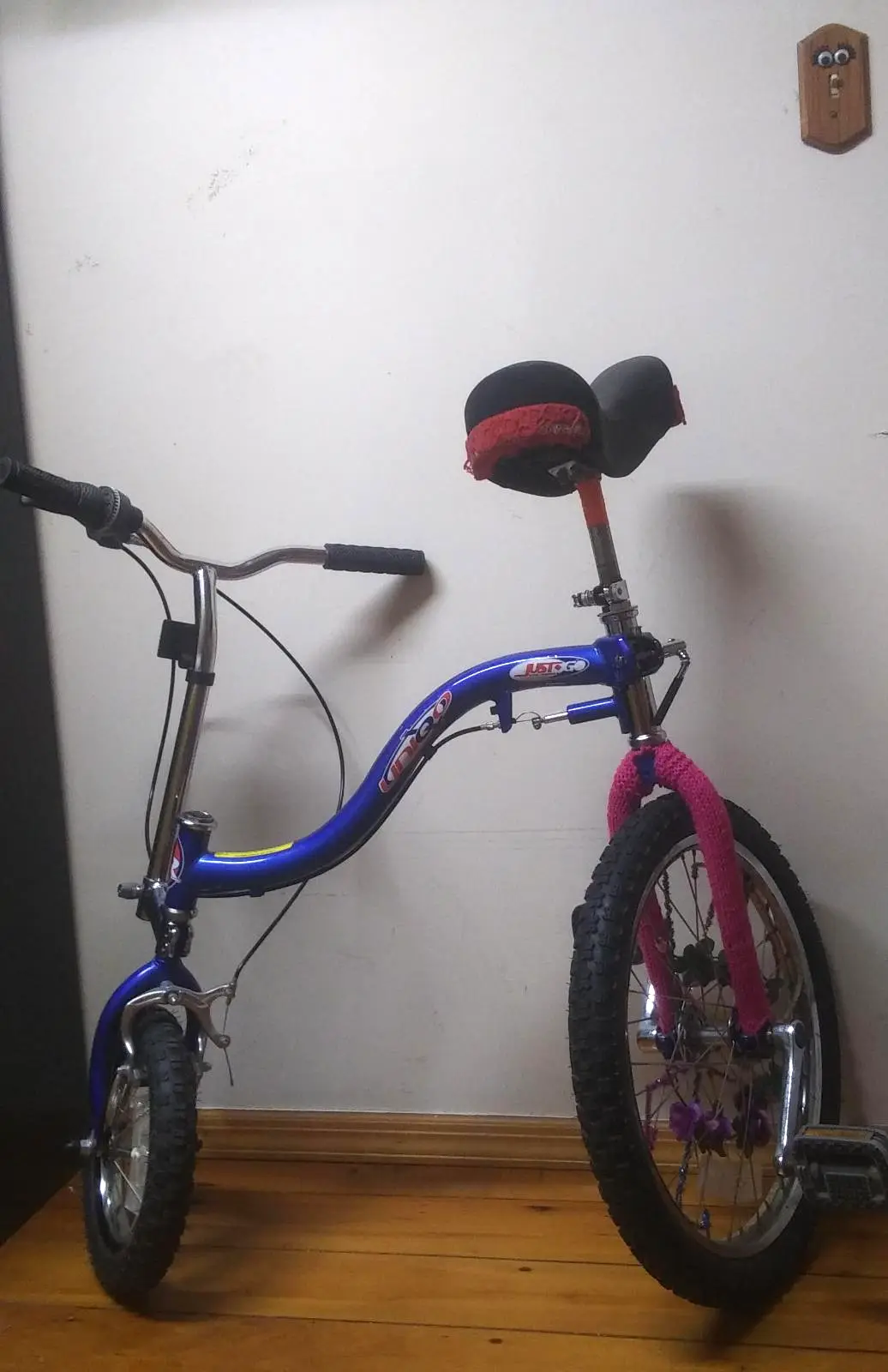 A blue 'Just Go' brand bicycle with a black seat wrapped in red fabric and pink knitted covers on the front fork, leaning…