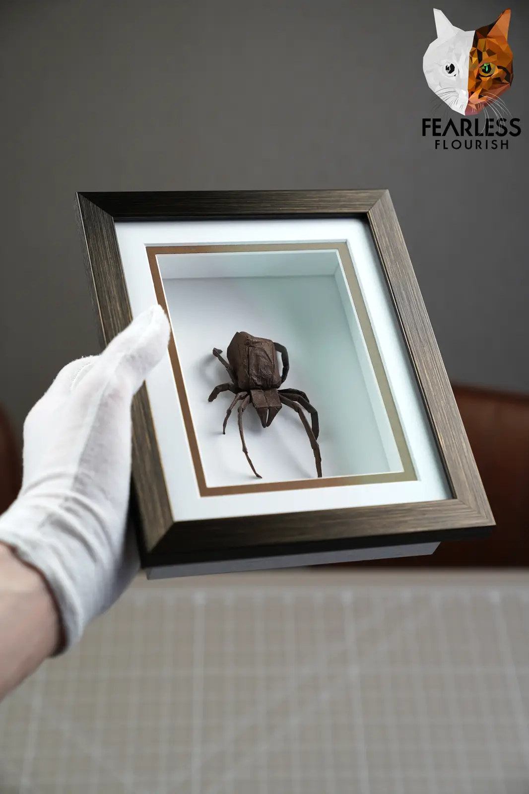 A gloved hand holds a shadowbox frame containing a meticulously crafted origami spider, presented with the logo 'FEARLESS…