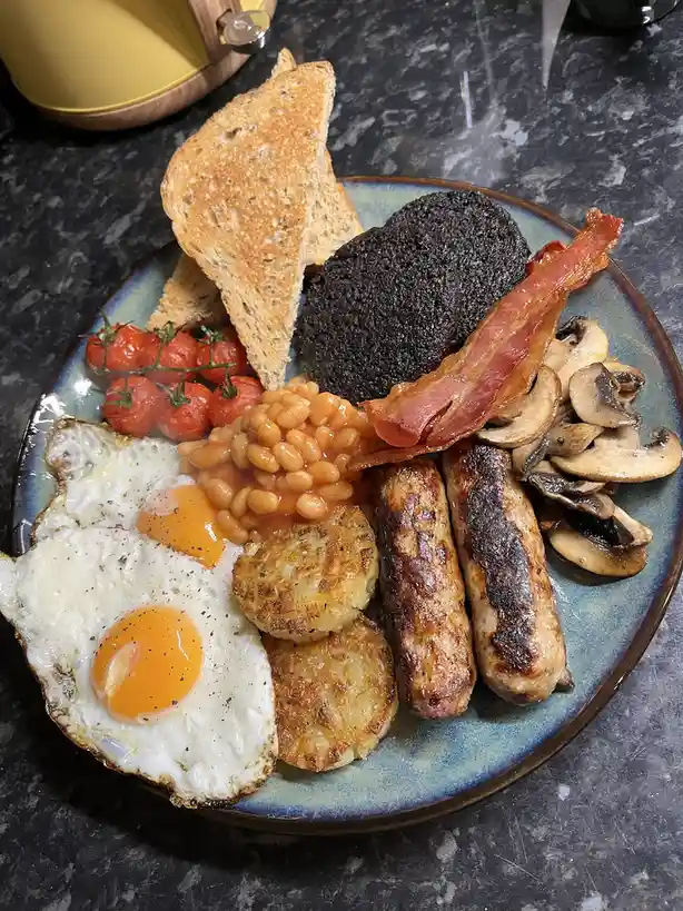 A plate of a full English breakfast featuring fried eggs, baked beans, sausages, bacon, black pudding, mushrooms, roasted…
