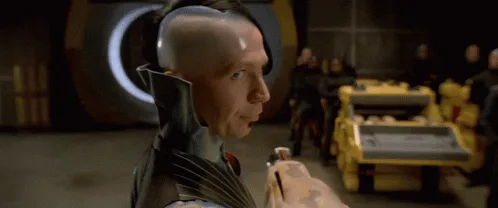 A bald man in a dark, high-collared outfit holds a small object while looking sideways with a skeptical expression. Behind…