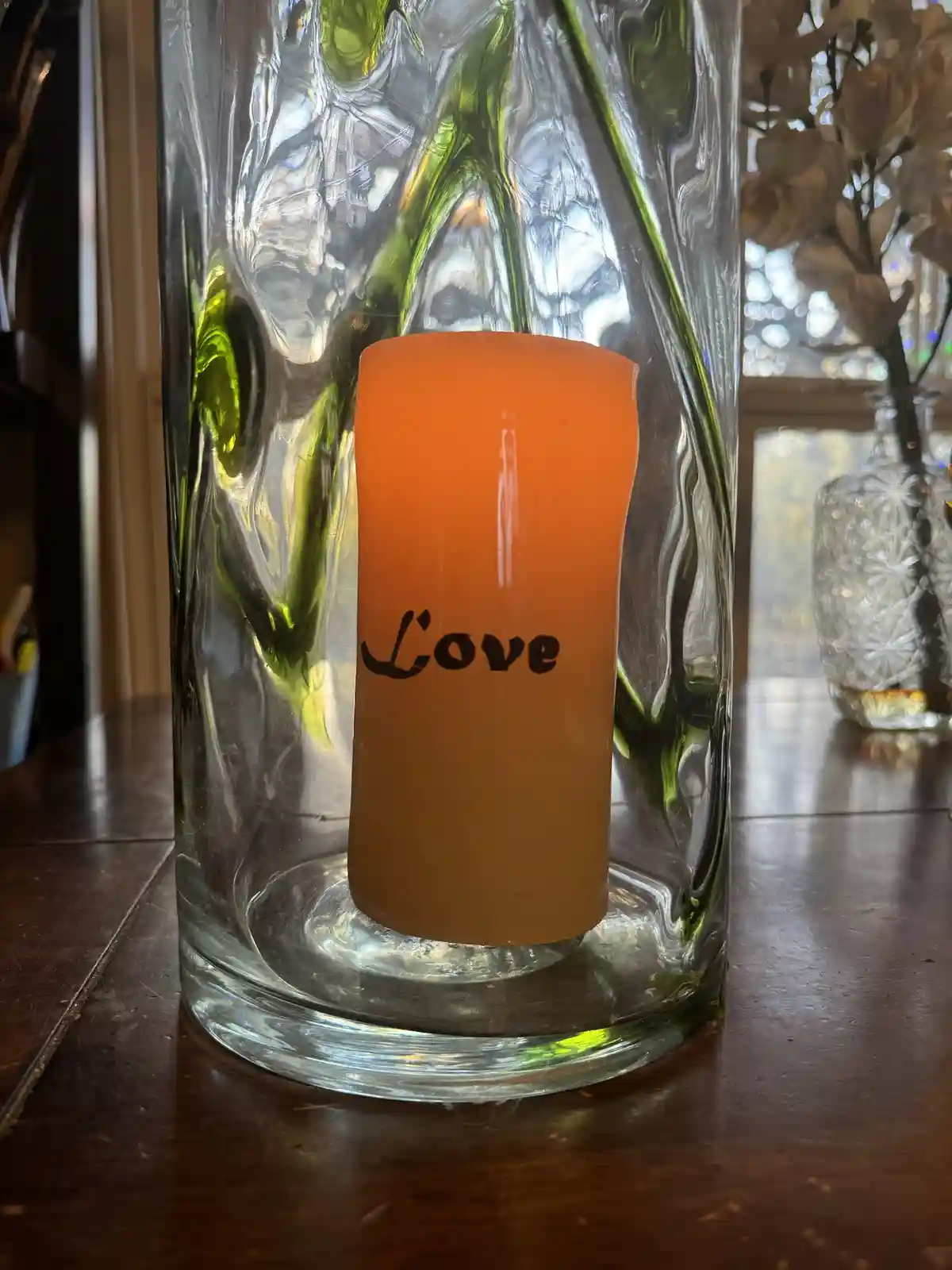 An orange, cylindrical candle with the word 'Love' written in black cursive font sits inside a tall, textured glass vase on…