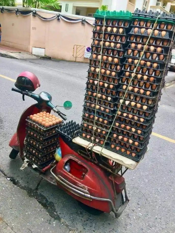 A red scooter is heavily overloaded with stacks of black egg cartons filled with brown eggs, strapped to the back and side…