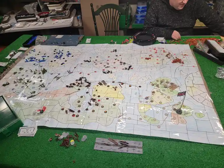 A person sits at a table playing a large-scale wargame with a hand-drawn world map, numerous miniature figurines, dice, and…