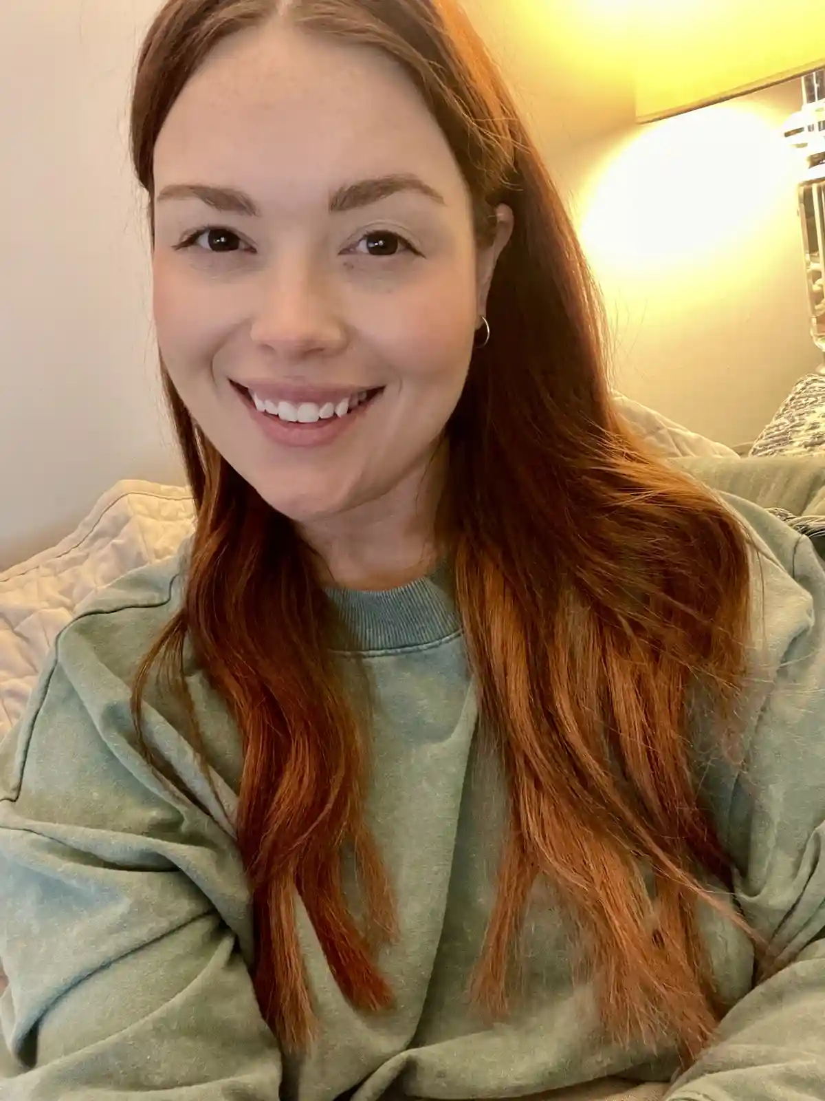 A woman with long reddish-brown hair and a wide, toothy smile is sitting on a couch wearing a light green sweatshirt. The…