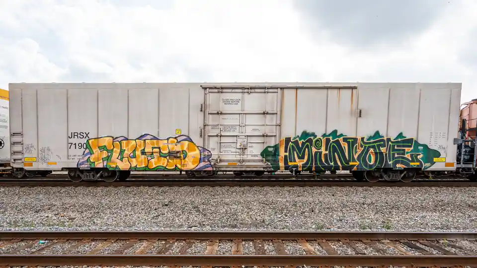 A side view of a white freight train car with graffiti on its sides, showing the words 'fizzed' and 'minutes' in stylized…
