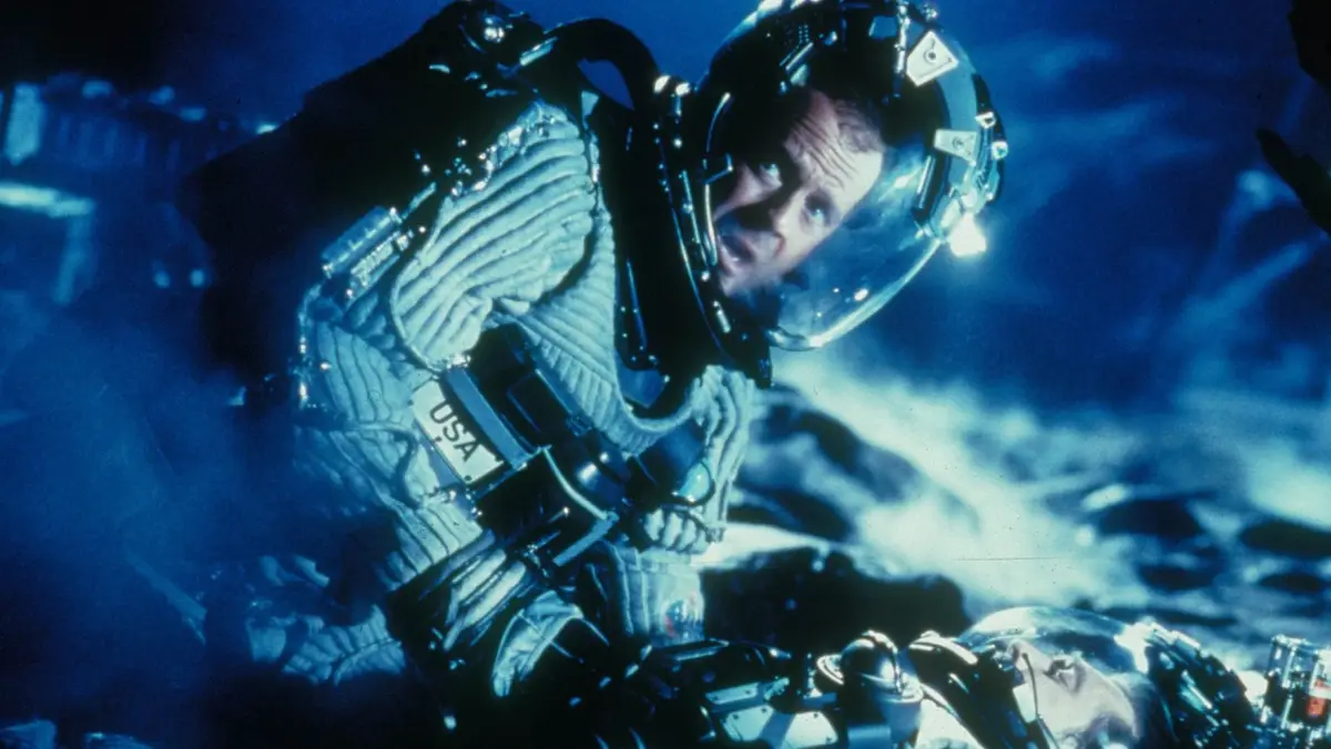 A man in a bulky space suit with a USA patch on his chest looks upward with a panicked expression, set against a blue, smoky…