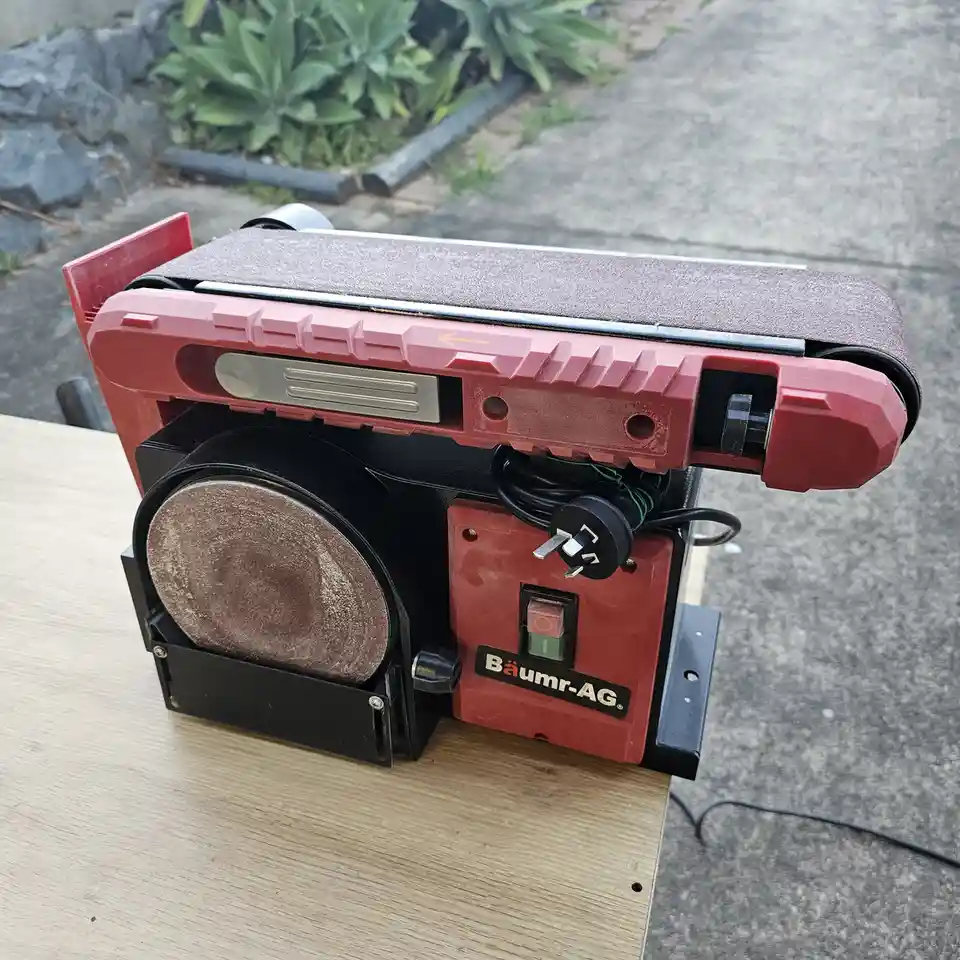 A red and black Baumer-AG belt and disc sander sits on a wooden table outdoors, with a worn sanding belt and a dusty disc…