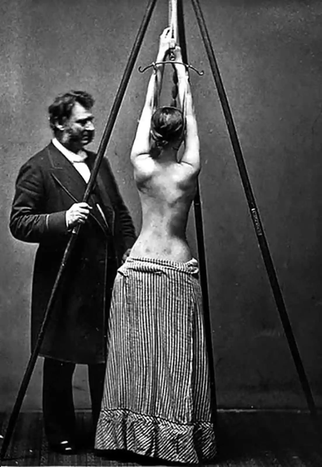 A vintage black-and-white photo shows a woman with her back to the camera, suspended upside down by her arms from a metal…