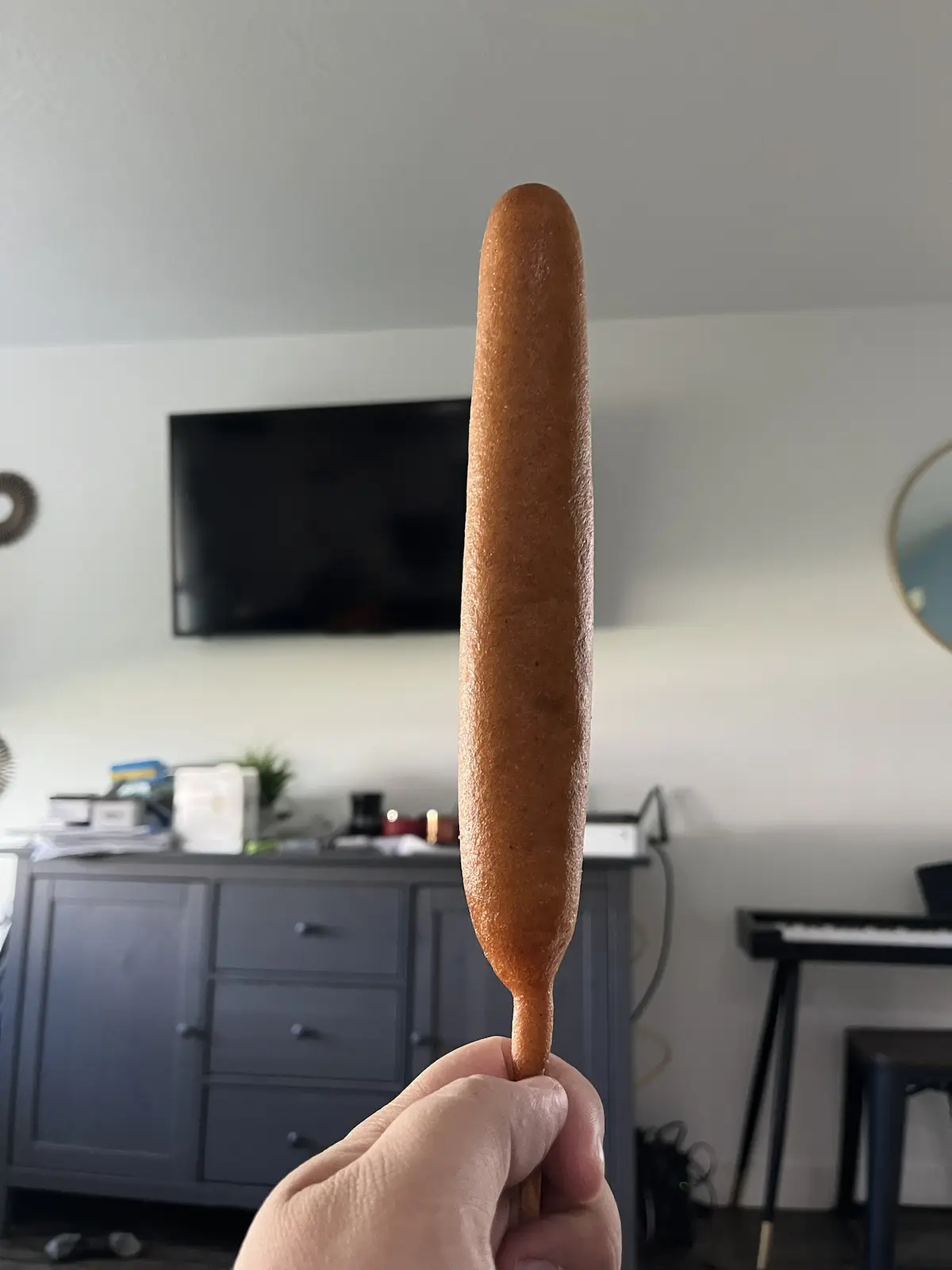 A hand holds up a long, golden-brown corn dog on a stick, centered in the frame against a blurred living room background…