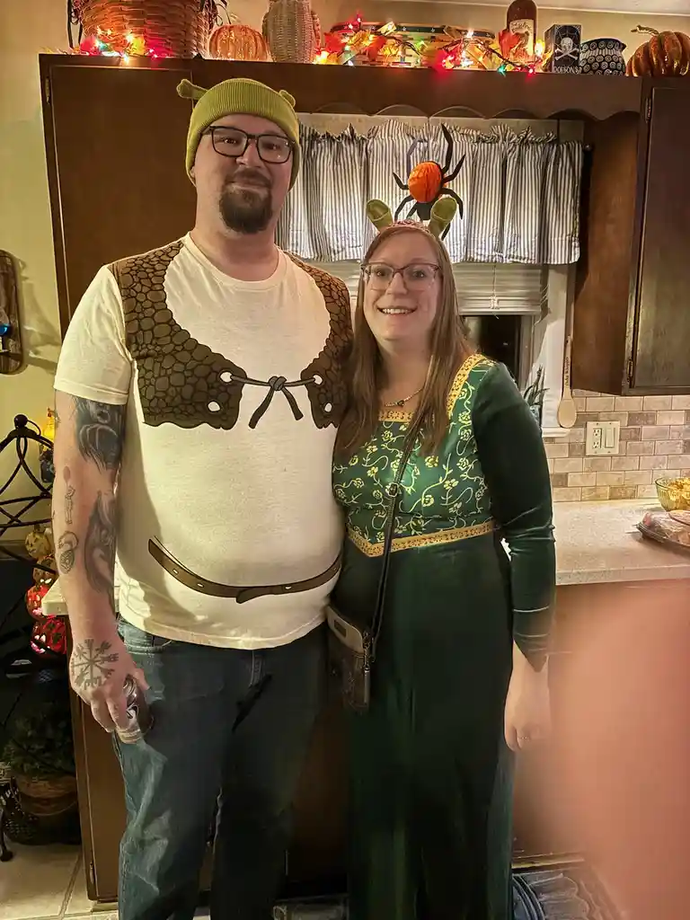 A man and woman dressed as Shrek and Donkey from the movie, standing in a kitchen decorated for Halloween. The man wears a…