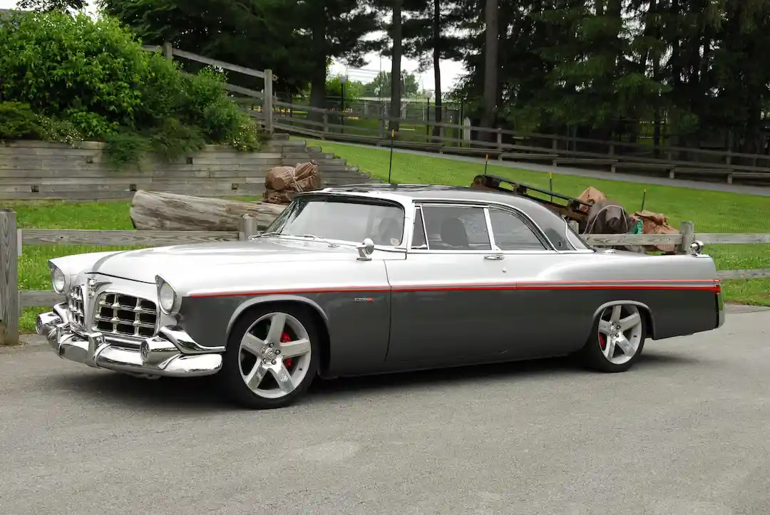 A silver and gray classic American car with modern custom wheels and a red pinstripe is parked on asphalt near a wooden…