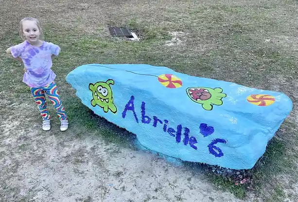A young girl in a tie-dye shirt and rainbow leggings stands proudly next to a large, light-blue painted rock decorated with…