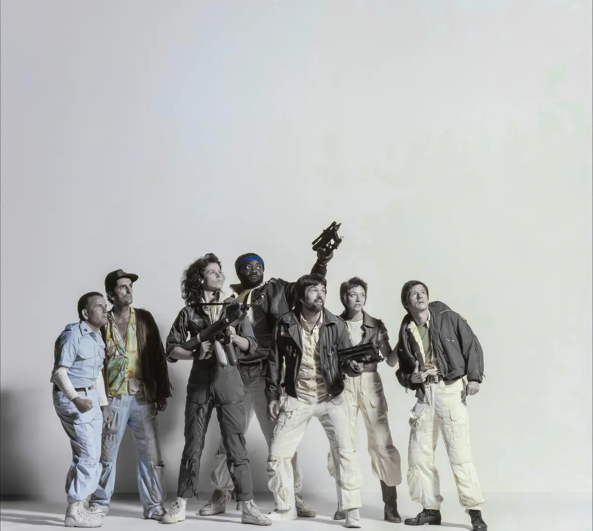 Seven people in sci-fi movie costumes, holding various weapons, all looking upward with expressions of awe or concern…