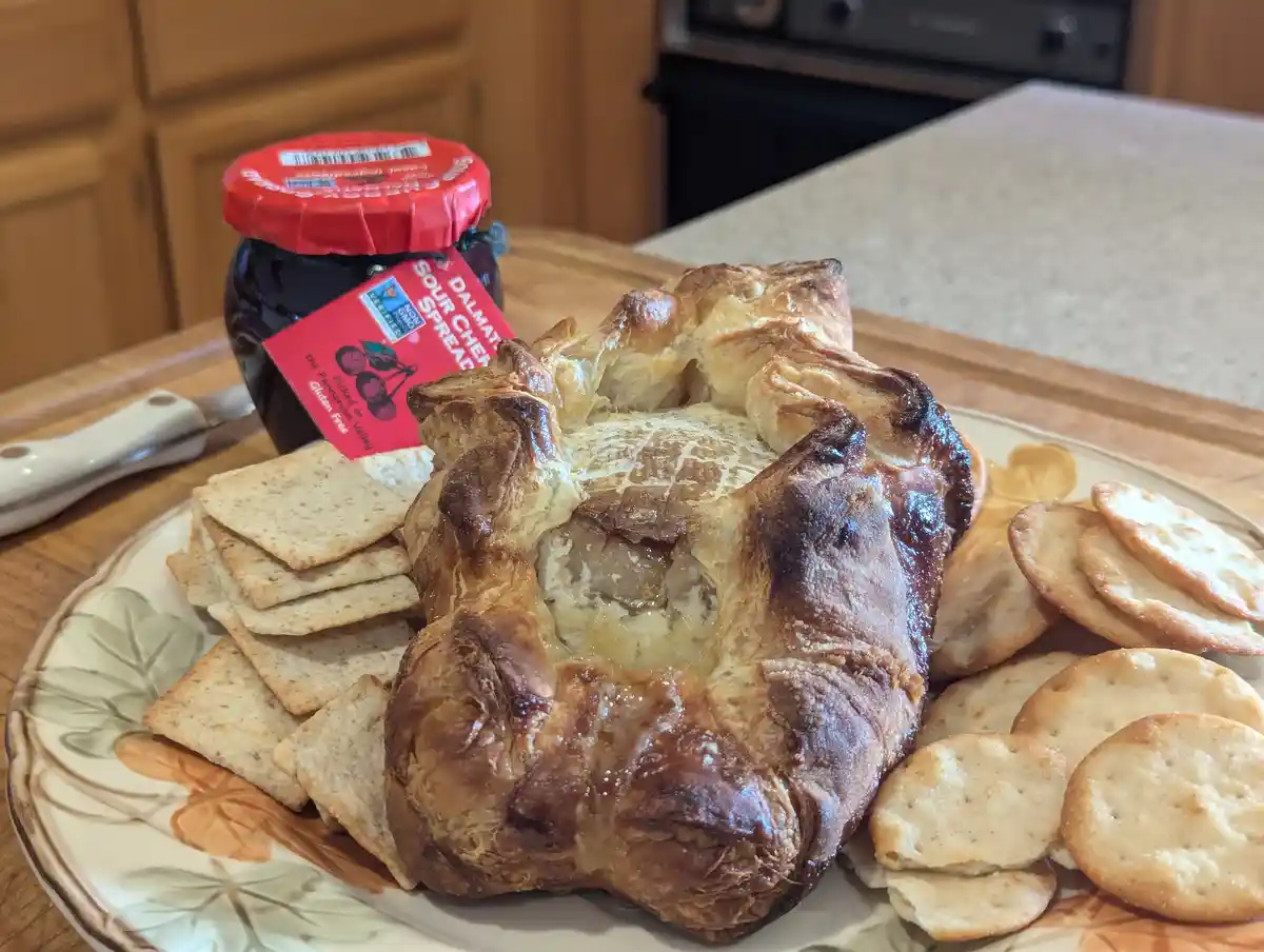 A baked cheese wheel with a golden-brown, flaky crust sits on a floral plate surrounded by square and round crackers. Behind…