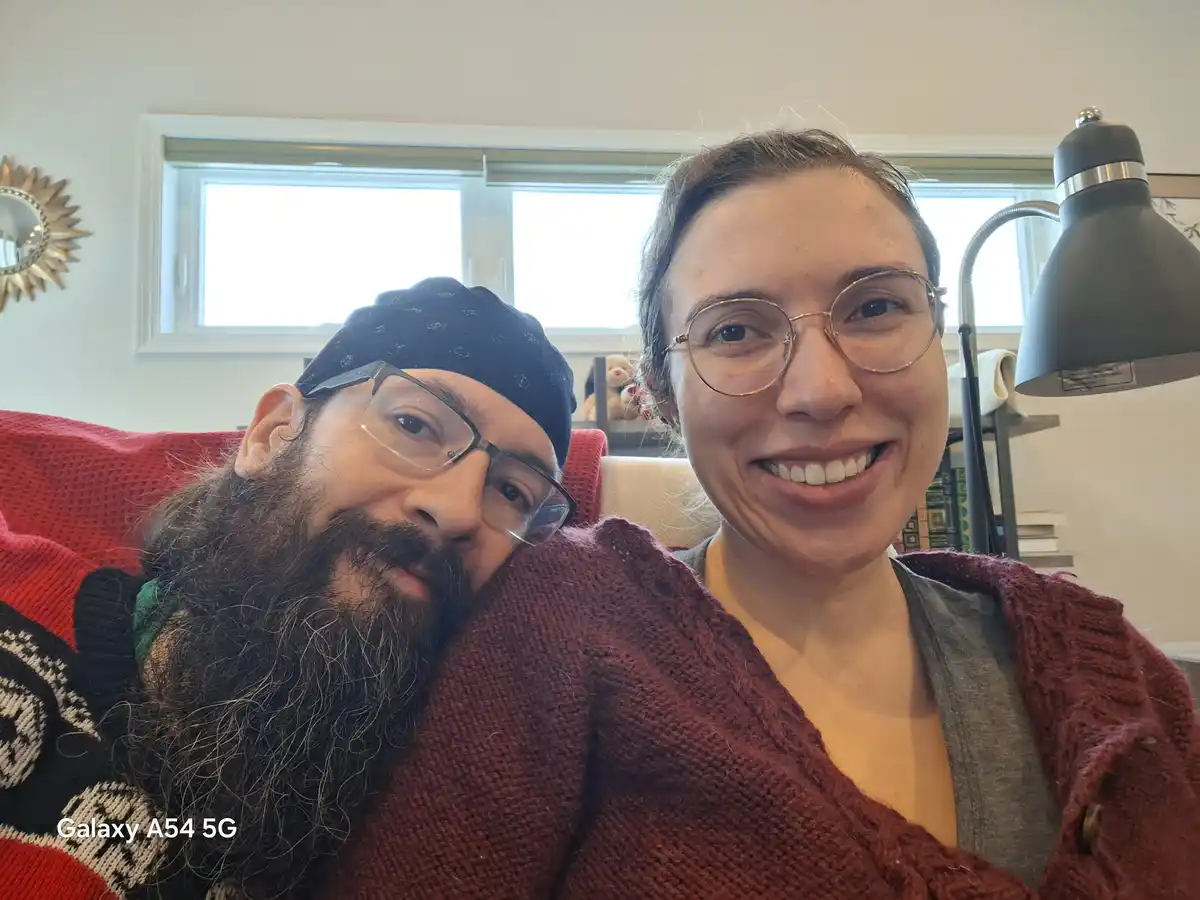 A selfie of a man with a long beard and glasses leaning on a woman with glasses, both smiling while sitting on a red couch…