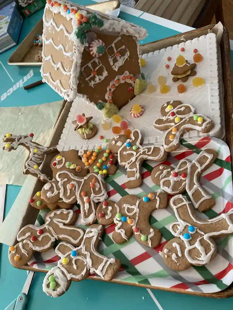 A photo shows a gingerbread house decorated with white icing and candy, alongside a tray full of gingerbread men decorated…