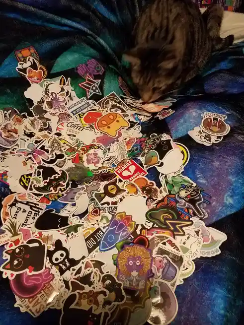 A tabby cat is seen sniffing or pawing through a large, colorful pile of assorted stickers scattered across a blue and…