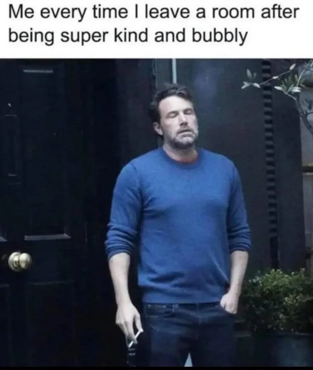 A man in a blue sweater stands outside a dark door, looking exhausted and drained, with one hand in his pocket and the other…