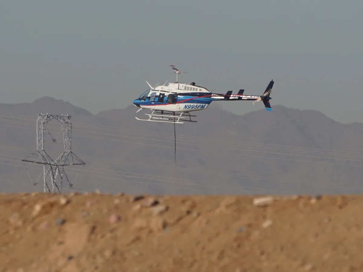 A white, blue, and red helicopter with registration N995PM is flying low over a desert landscape with mountains in the…