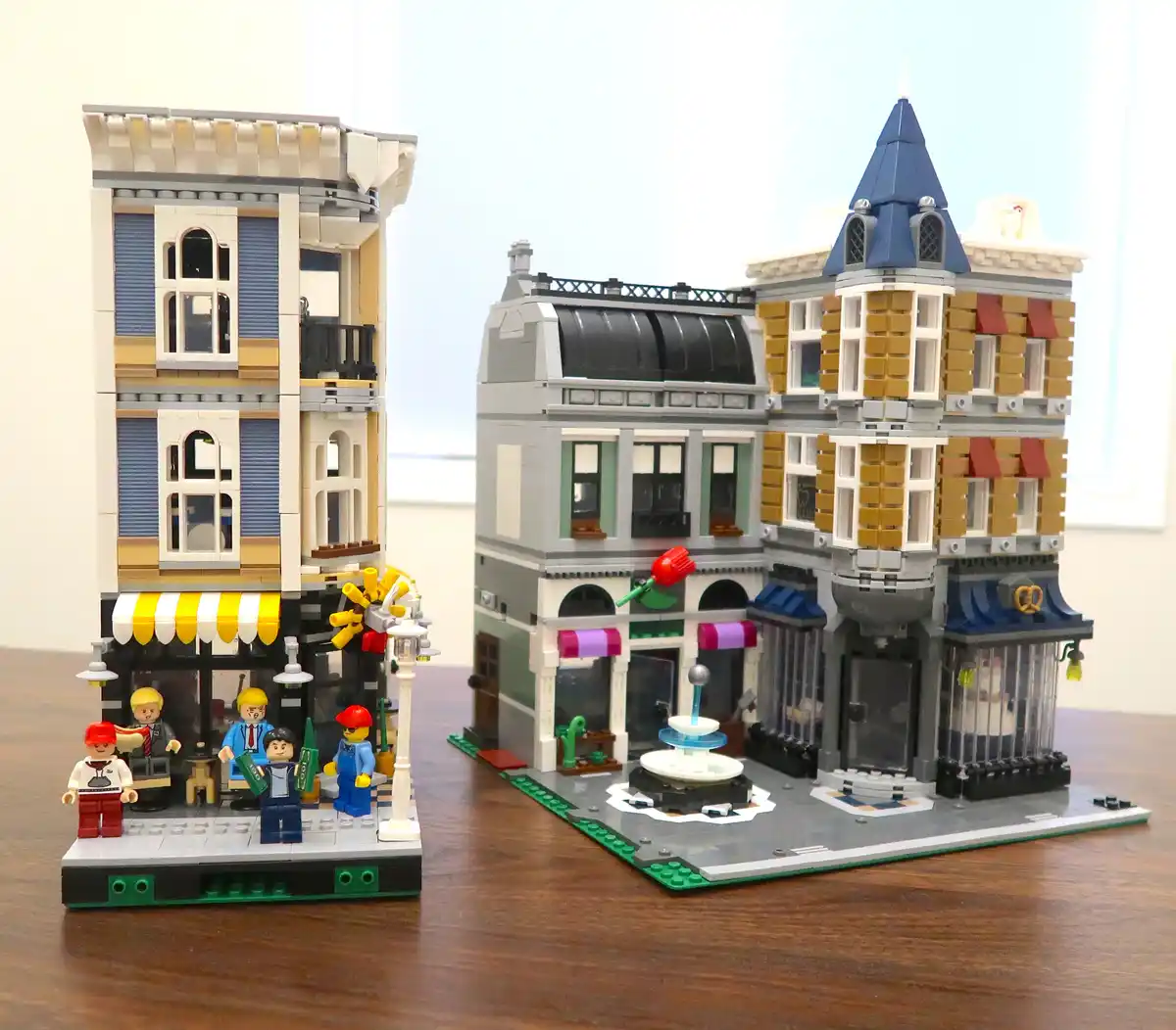 Two detailed LEGO city buildings sit side-by-side on a wooden surface, one with a yellow-and-white striped awning and…