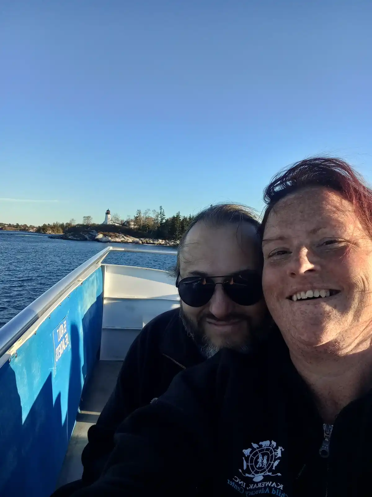 A selfie of two smiling people on a boat with a lighthouse visible in the distance under a clear blue sky. The boat has a…
