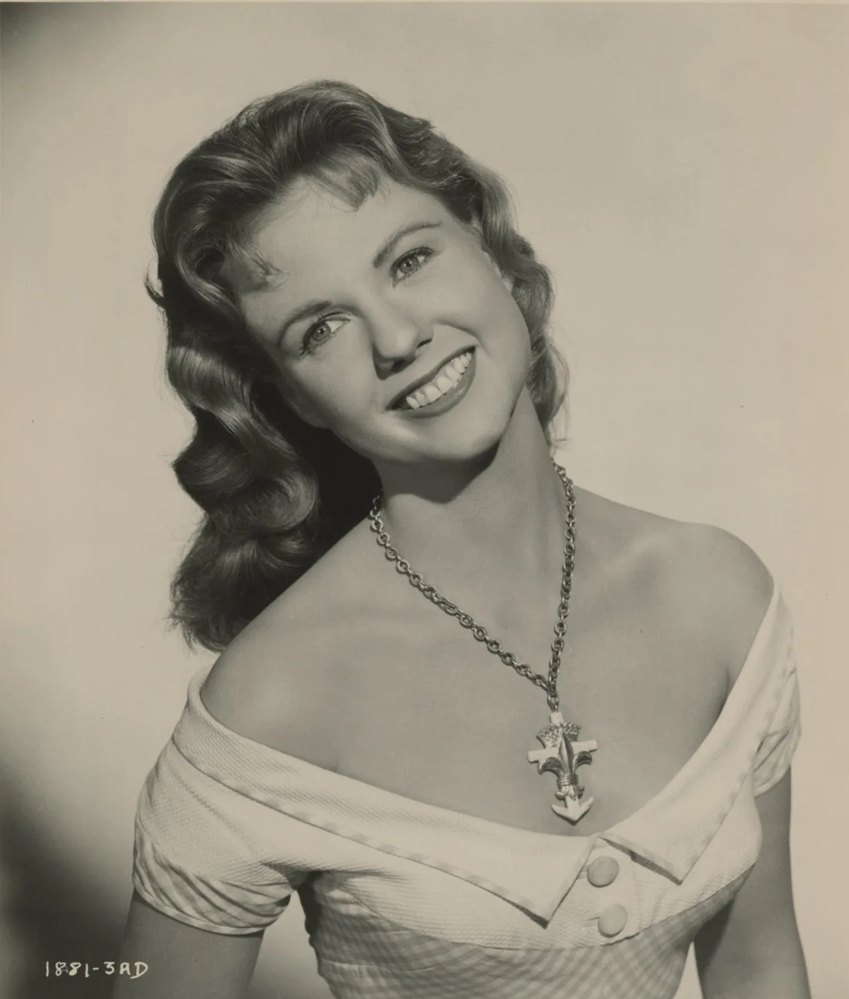 A vintage black-and-white studio portrait of a smiling woman with styled curls, wearing an off-the-shoulder dress and a…