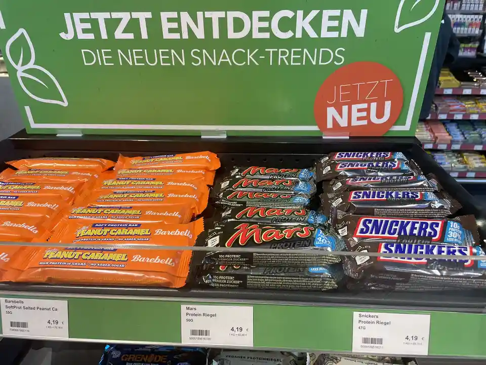 A supermarket shelf displays protein bars, including Barebells, Mars, and Snickers Protein Riegel, under a green sign that…