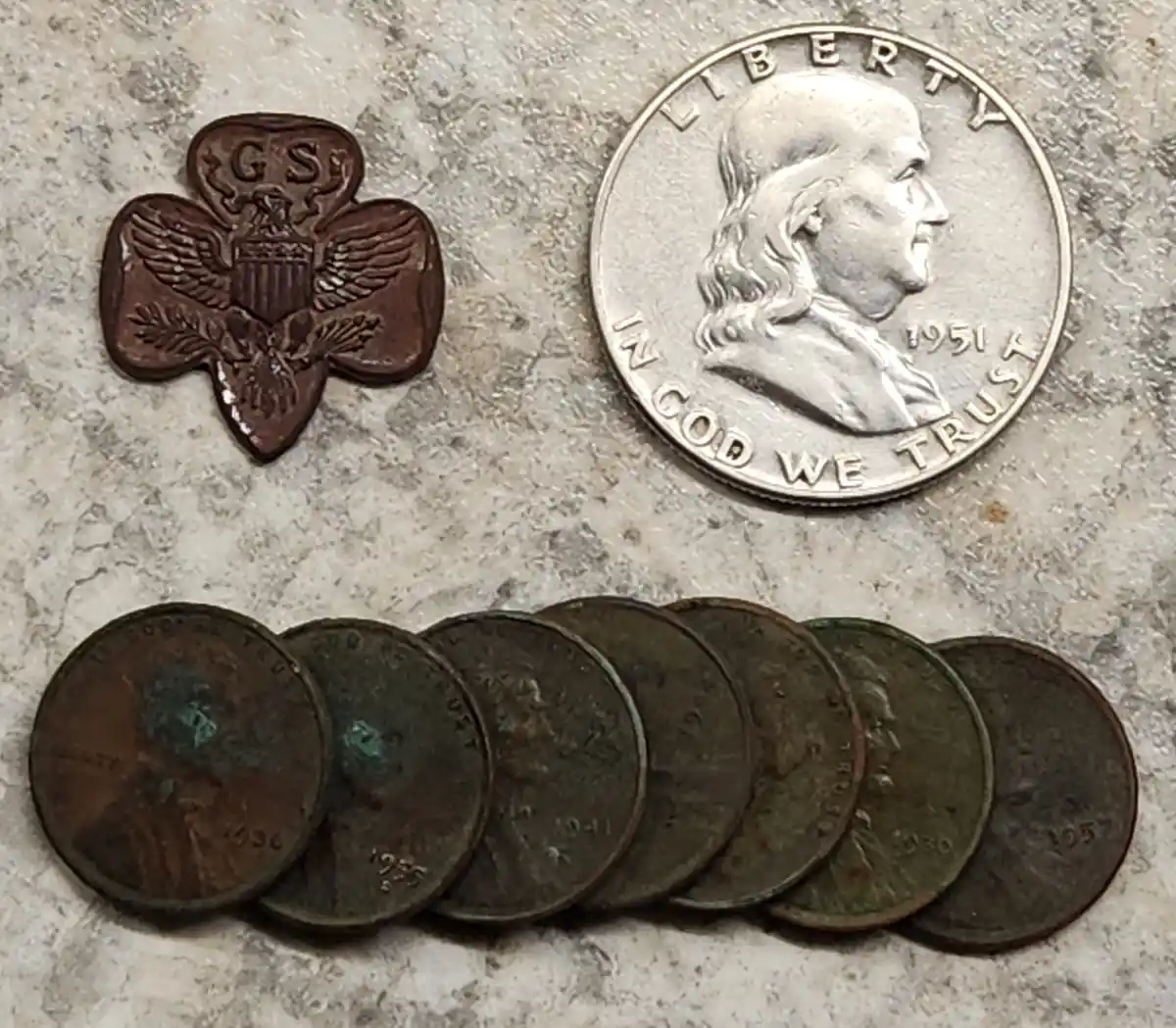 An image displays a Girl Scout trefoil pin next to a 1951 U.S. quarter and a row of seven old, tarnished U.S. pennies. The…