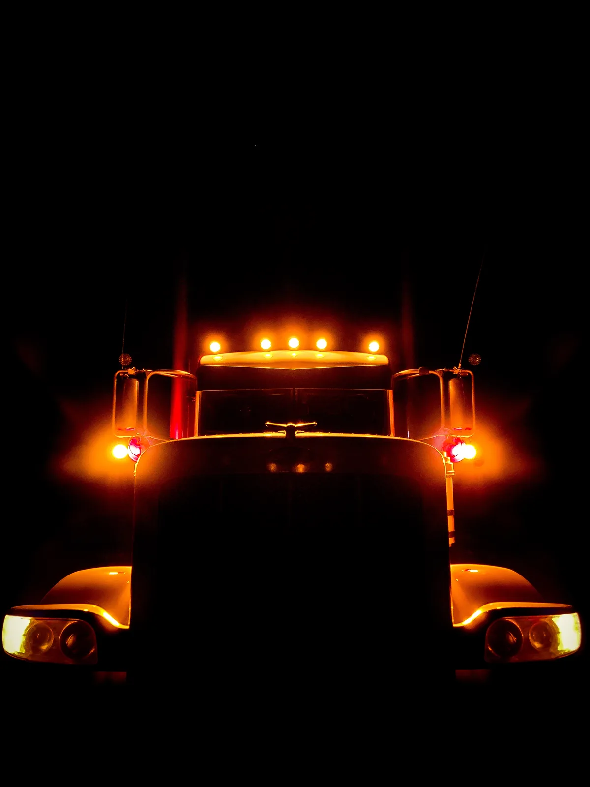 A front-facing shot of a large semi-truck at night, its numerous orange and white lights glowing brightly against a…