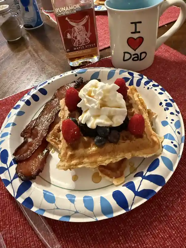 A plate of waffles topped with whipped cream, raspberries, and blueberries sits next to crispy bacon. Behind it is a mug…