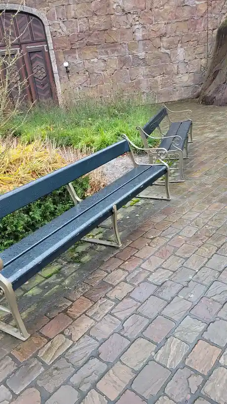 Two empty, dark-colored park benches with ornate metal frames sit on a wet, stone-paved path, angled slightly away from each…