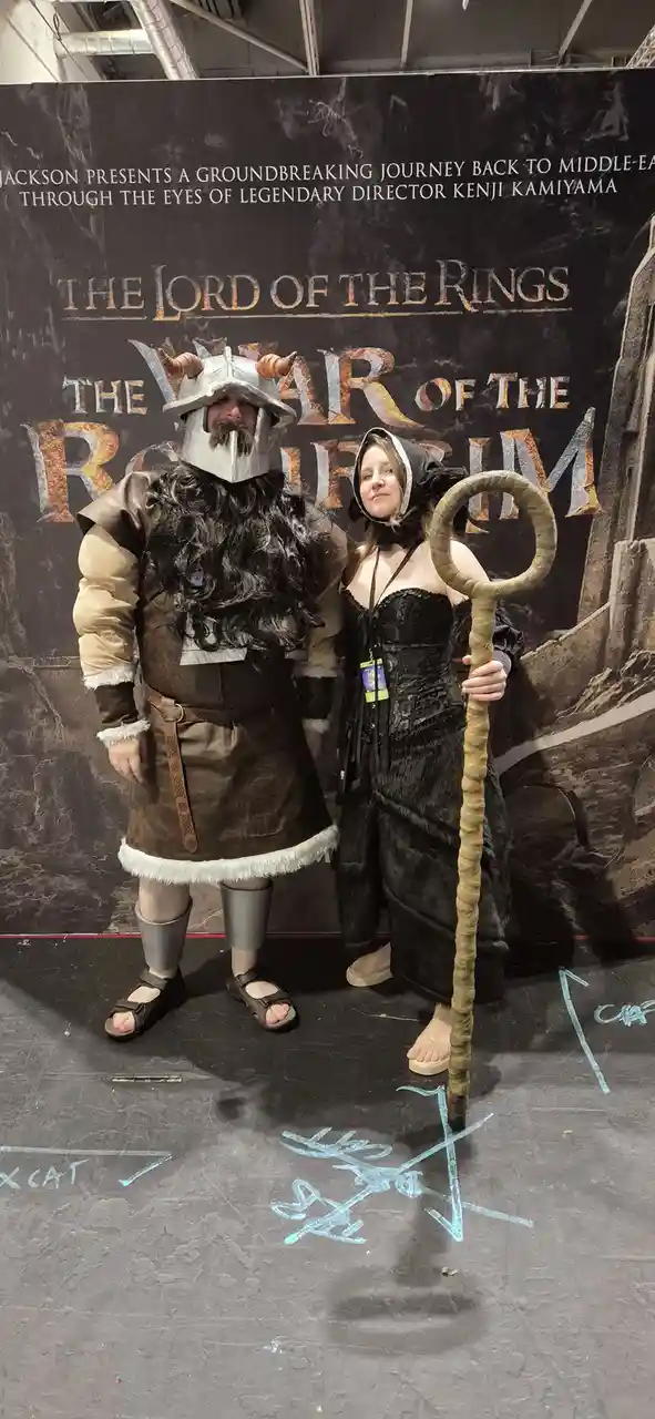 Two people in Lord of the Rings cosplay stand before a promotional backdrop for 'The Lord of the Rings: The War of the…