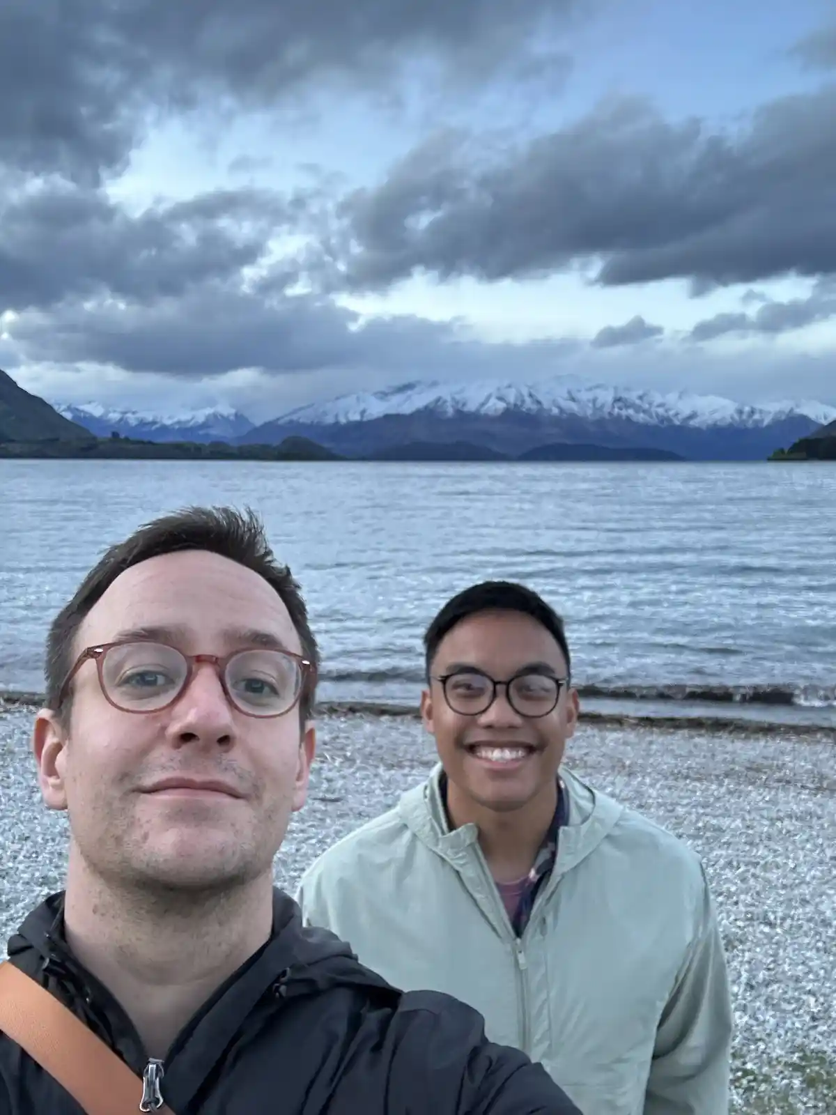 Two men stand on a pebbled lakeshore with snow-capped mountains in the background under a cloudy sky; the man in the…