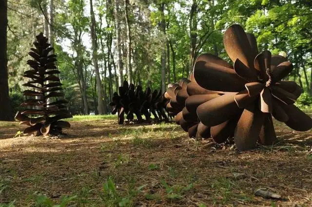 A row of large, rusted metal sculptures shaped like pine cones sits on forest floor, with the closest one appearing to be…