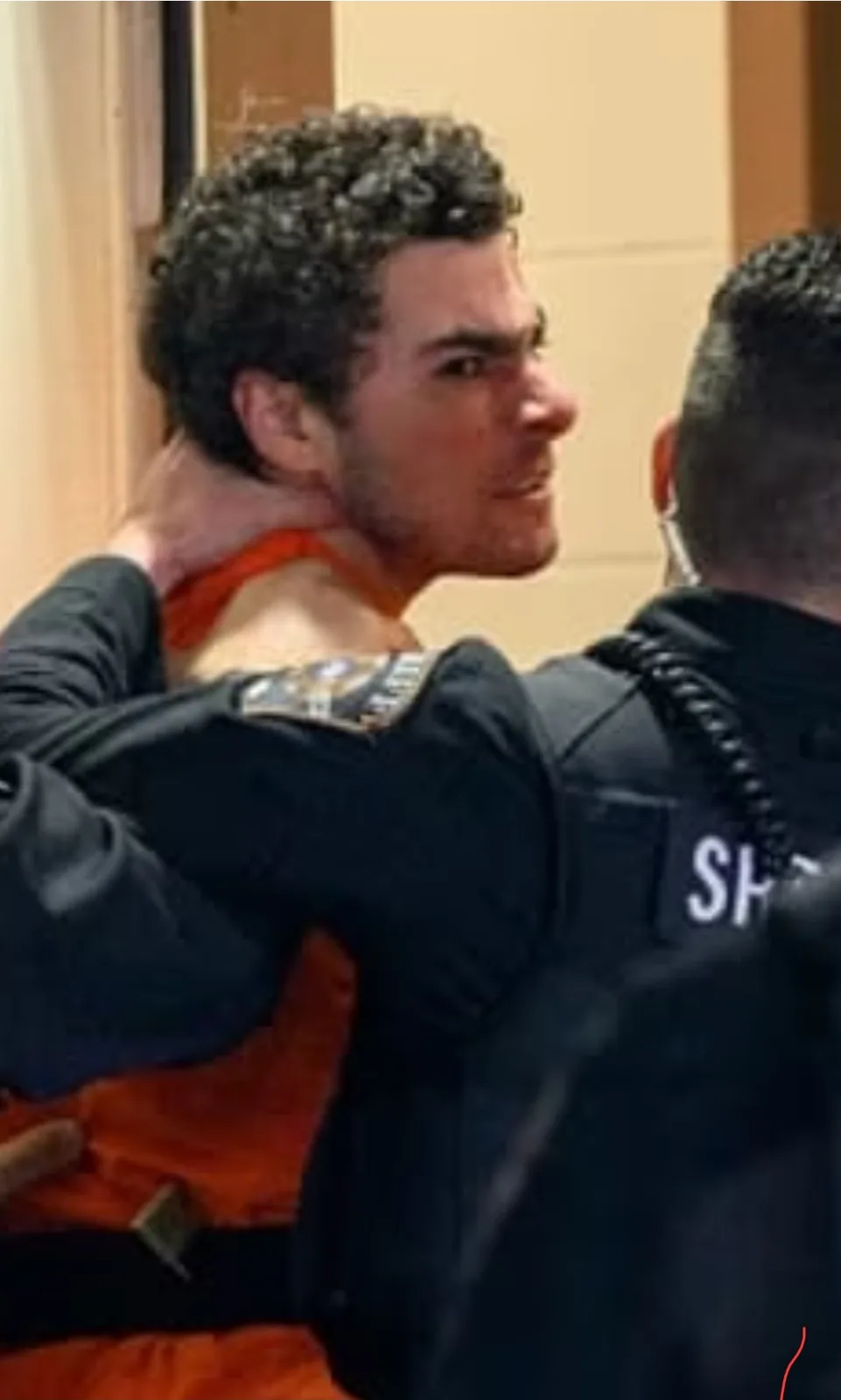 A man in an orange jumpsuit is being restrained by a police officer whose hand is around his neck. The officer's uniform has…