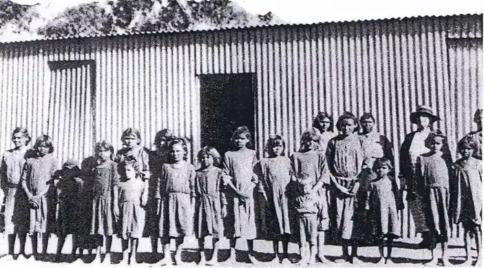 A black-and-white historical photo shows a group of children and adults standing in front of a corrugated metal building…