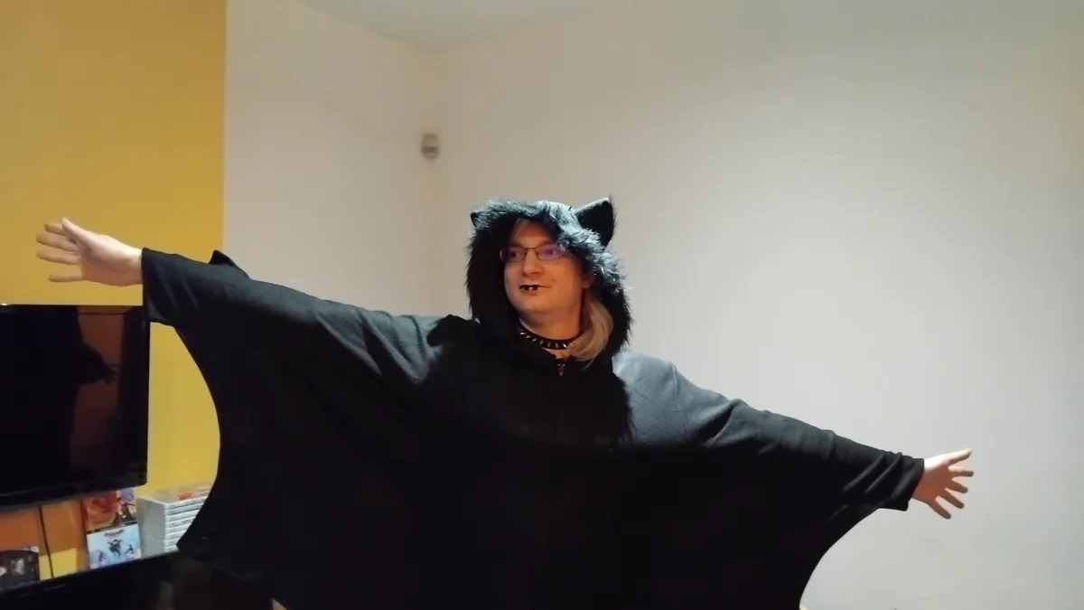 A person wearing a black, furry bat costume with cat ears and fangs, arms spread wide in a room with yellow and white walls…
