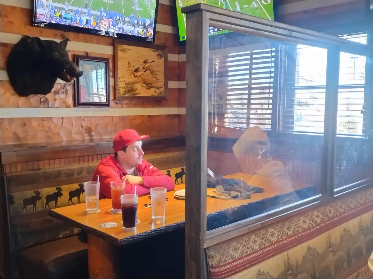 A man in a red hoodie and cap sits at a wooden booth in a rustic sports bar, looking toward a glass partition. Behind the…
