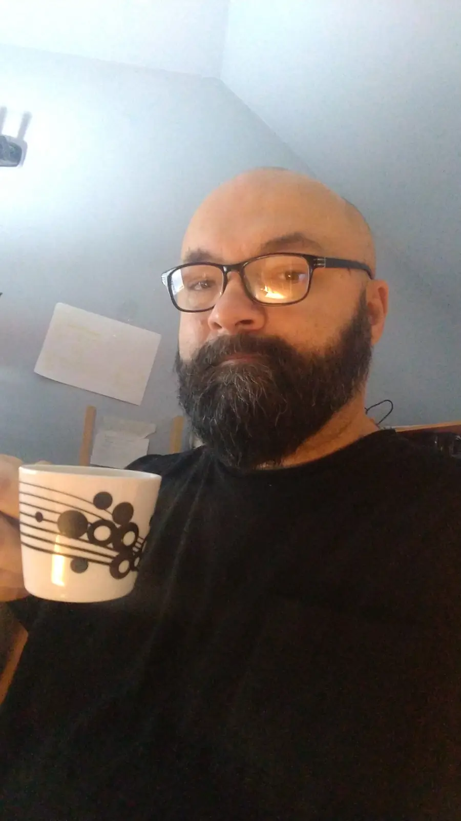 A bald man with a full beard and glasses holds a white mug decorated with black musical notes and circles. He looks directly…
